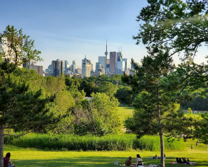 Riverdale Park in Toronto people sitting under lush trees and greenery with picnic blankets enjoying each other's company with the Toronto skyline in the background taken by James Thomas