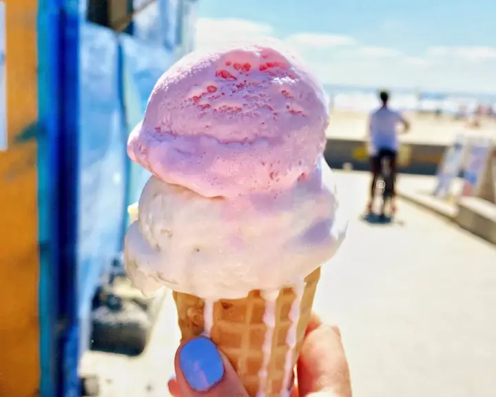 two scoop of what looks like vanilla and strawberry ice cream slightly melting in front of what looks like a food truck with a person on a bike in the background and the beach in the background of them by GG Lemere. Ice cream and other food can be bought at Woodbine Beach