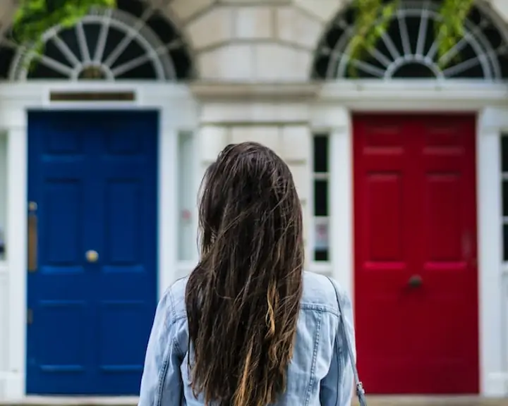 person with hip length hair and a jean jacket on with back towards camera standing in front of two doors - one is red - one is blue - seems like a play on the Matrix
