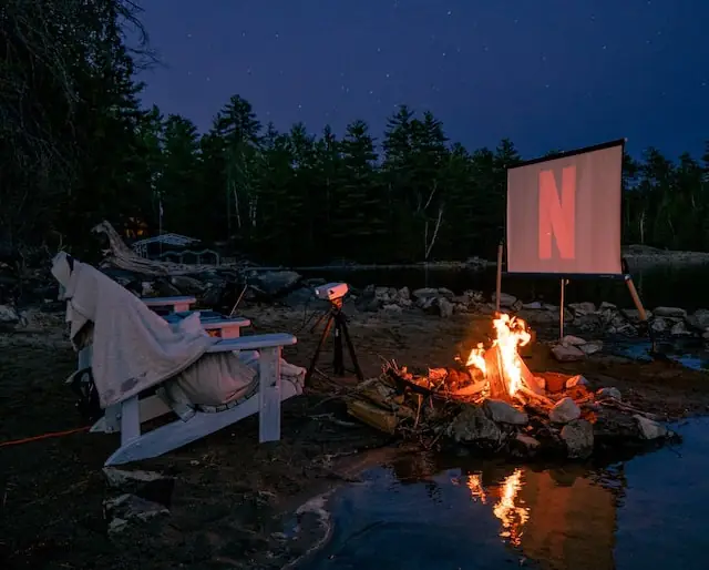 outdoor movie with muskoka chairs and fire next to a body of water and woods