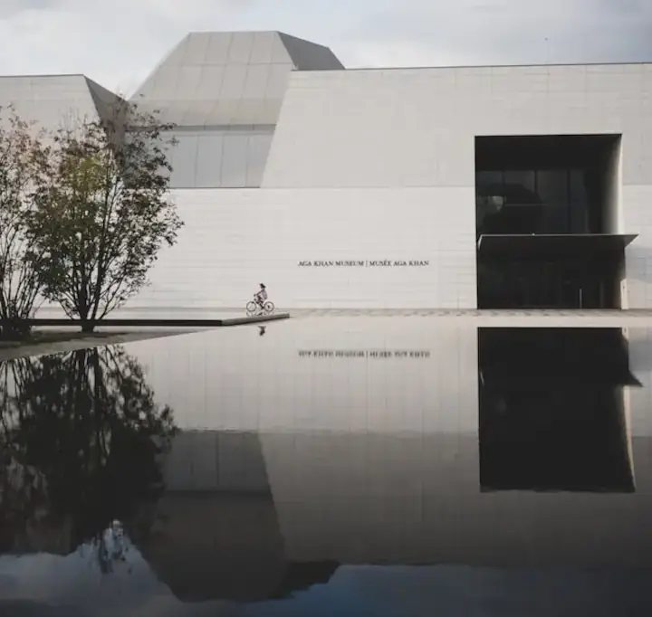 Aga Khan Museum in Toronto with reflection pool in front and a person with long hair and wearing a dress biking in front