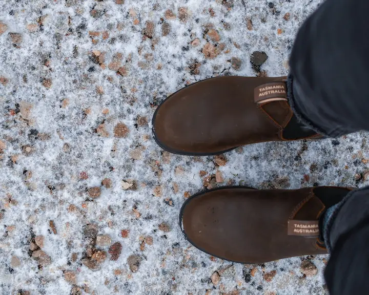 person wearing a pair of brown Bludstone boots with the Australia Tasmania tag clearly showing stepping on some snow
                    covered pebble path