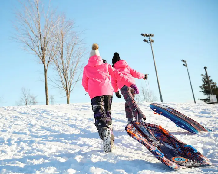 Two people walking up a snowy hill in pink winter jackets and snow pants toboggans in hand