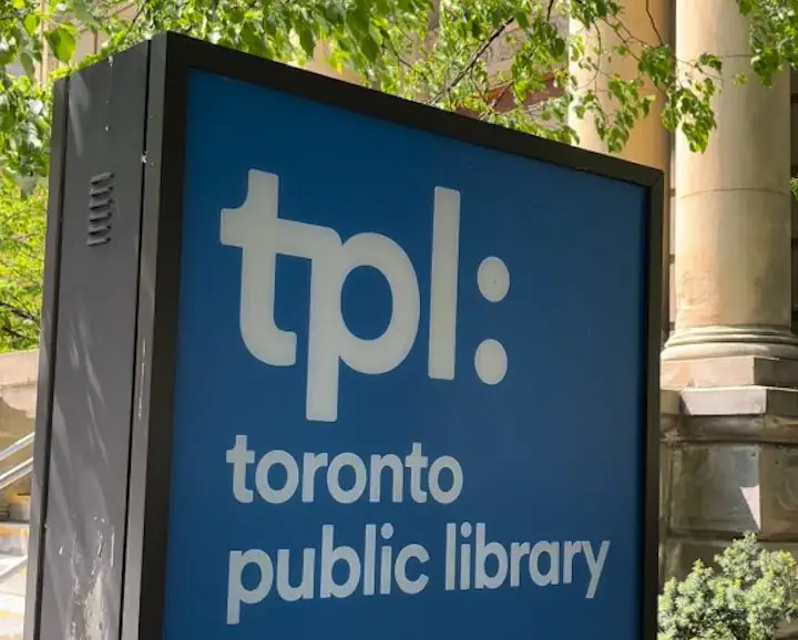 Toronto Public Library sign in blue and white TPL with columns in the background and green tree leaves above